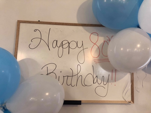 Happy 80th Birthday written on a whiteboard surrounded by blue and white balloons